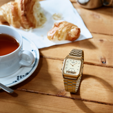 Gold watch on a wooden table with a cup of tea and croissants.
