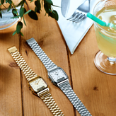 Two watches, one gold and one silver, on a wooden surface with drinks and cutlery in the background.