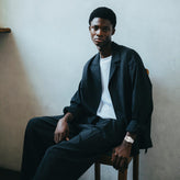 Man sitting on a chair wearing a dark jacket and white shirt against a plain wall.