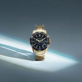 A gold-tone stainless steel chronograph watch with a black dial and bezel, displayed on a dark gray surface with dramatic spotlighting.