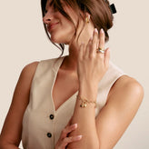 A woman wearing a delicate gold chain bracelet with small charms on her wrist, paired with gold hoop earrings and stacked gold rings on her finger, set against a solid beige background.