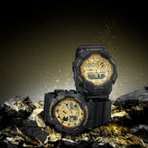 Two black Casio G-Shock digital sports watches with gold-toned dials displayed on dark rocks against a dramatic dark background with gold light effects.